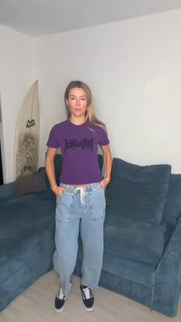 Violets pieguloša T-krekls ar melnu grafisko dizainu, kopā ar brīviem gaisīgi ziliem džinsa biksēm un melnām sporta kurpēm, stāvot uz zilas dīvāna.