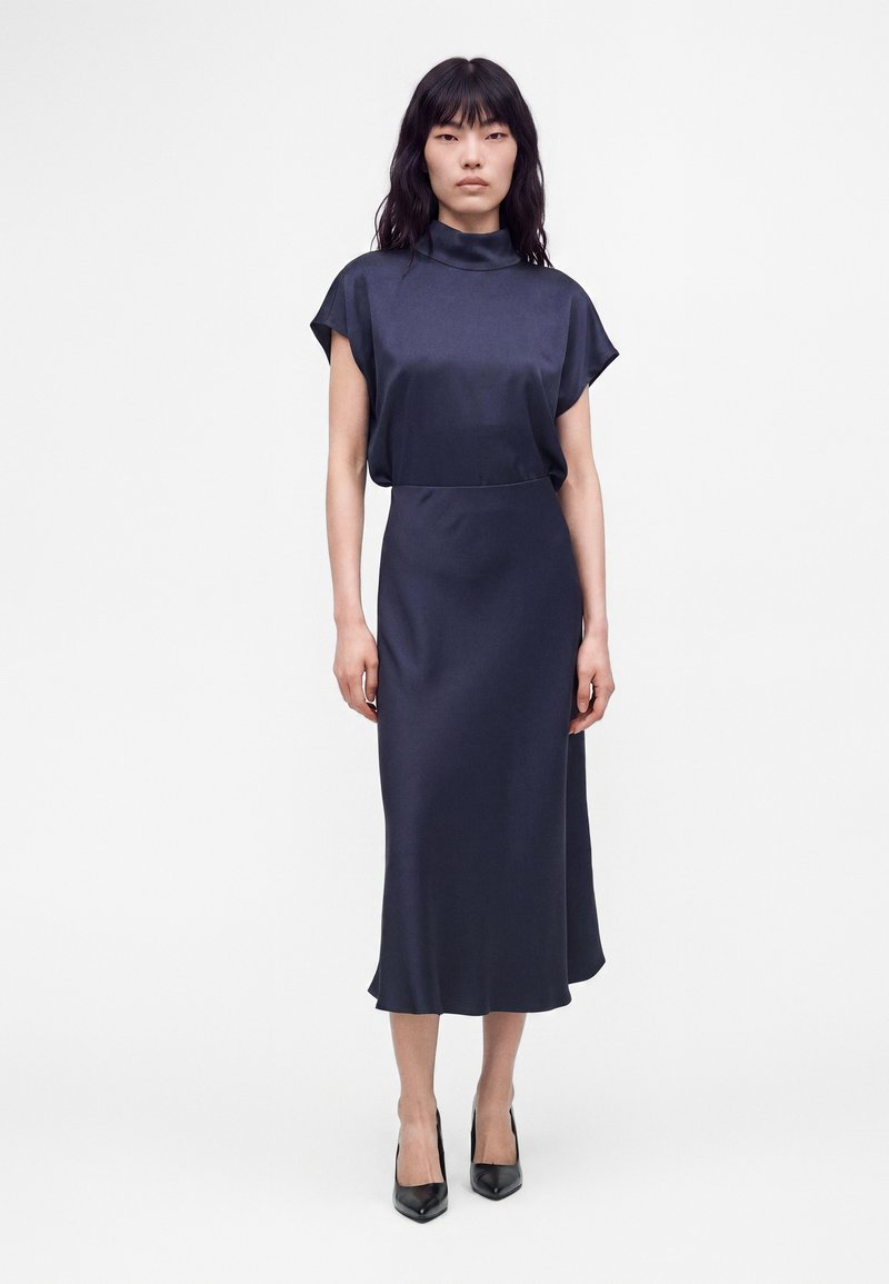 Femme debout sur fond blanc portant un haut en satin bleu marine à col montant et une jupe midi assortie avec des talons pointus noirs.