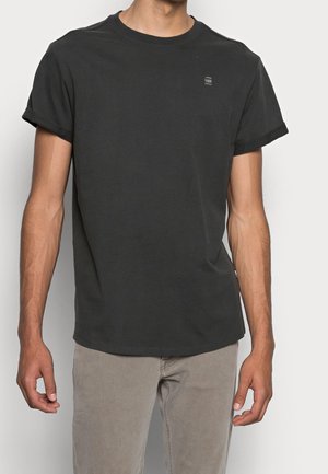 Mand iført en ensfarvet sort kortærmet T-shirt med opkrøllede ærmer og lysegrå bukser, stående foran en hvid baggrund.