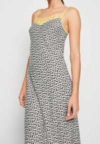 Robe noire sans manches avec un motif floral blanc, présentant une bordure en dentelle jaune le long du décolleté et des bretelles ajustables.