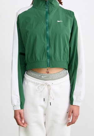 Personne portant une veste zippée Nike verte et blanche, un pantalon de survêtement blanc à cordon, une ceinture Nike visible et un piercing au nombril.