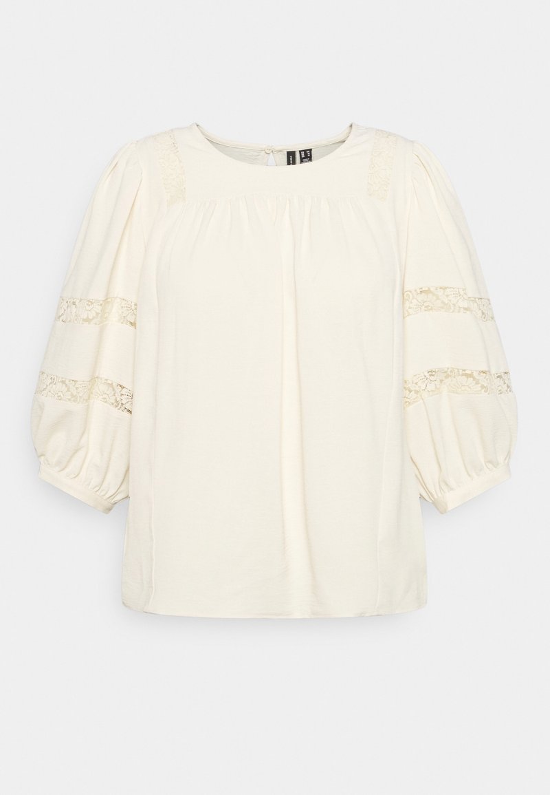 Blouse crème avec des manches courtes et bouffantes. Présente des détails en dentelle sur les manches et les épaules, avec des fronces au niveau du col. Tissu doux.