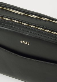 Black leather bag with gold zippers, featuring a prominent gold "BOSS" logo. Smooth texture and sleek design, ideal for various uses.