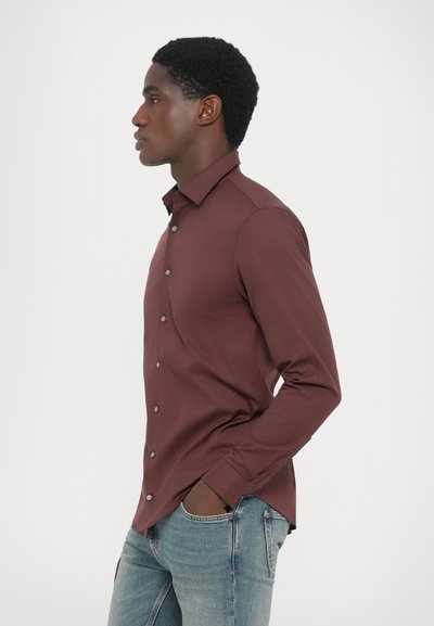 Camisa de botón en color marrón con un corte ajustado, que presenta un collar estándar y botones grises. Combinada con jeans de denim azul.