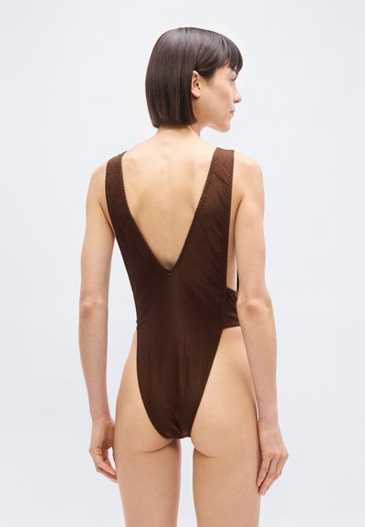 Γυναίκα με κοντά καστανά μαλλιά που φοράει καφέ ολόσωμο μαγιό με βαθύ V στην πλάτη, στέκεται μπροστά σε απλό φωτεινό φόντο.