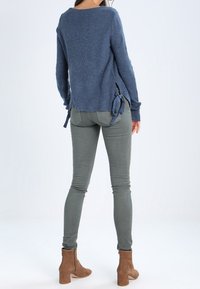 Maglione azzurro lavorato a maglia con lacci laterali, abbinato a jeans grigi slim-fit e stivaletti color cammello, visto da dietro.