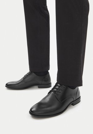 Scarpe eleganti nere stringate in pelle indossate con pantaloni neri su sfondo bianco, con un primo piano di piedi e gambe inferiori.