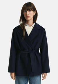 Marine jas gemaakt van een wolmix, met een ceintuur in de taille, gelapen revers en brede mouwen. Gecombineerd met een lichte trui en een colbert.