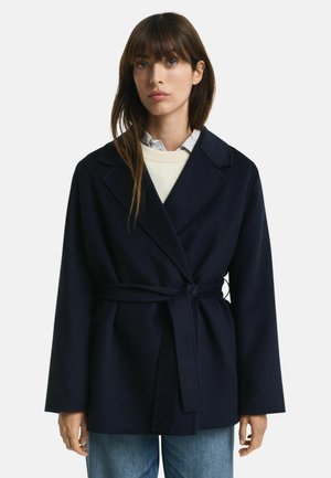 Navy coat made of wool blend, featuring a belted waist, notched lapels, and wide sleeves. Worn over a light sweater and collared shirt.