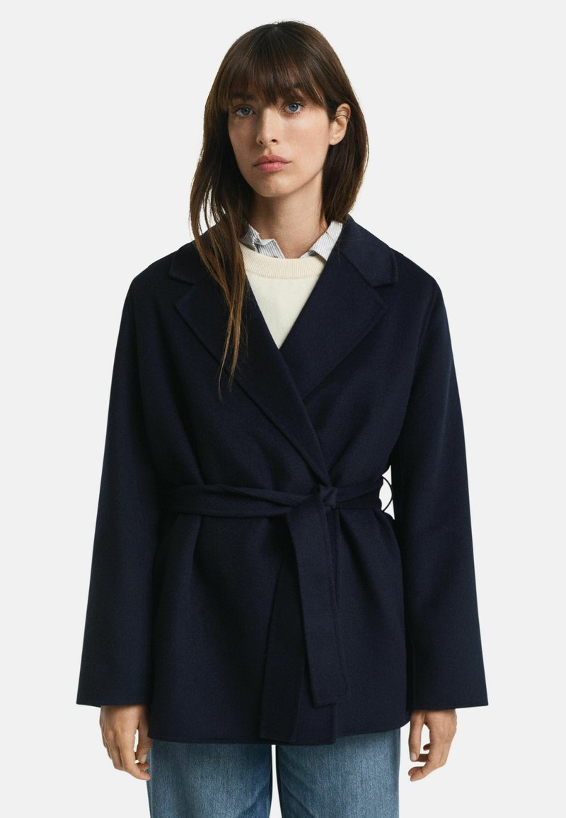 Marine jas gemaakt van een wolmix, met een ceintuur in de taille, gelapen revers en brede mouwen. Gecombineerd met een lichte trui en een colbert.