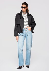 Femme portant une veste noire courte, une chemise blanche rayée, un jean taille haute bleu clair, des talons noirs et des lunettes de soleil cat-eye, debout avec une main dans la poche.