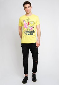Yellow T-shirt featuring cartoon characters, text "IT FEELS NICE TO DO GOOD". Paired with black ripped jeans and black shoes.