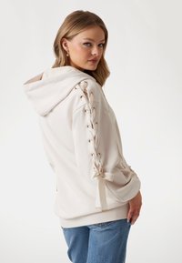 Cream hoodie with lace-up details on sleeves, satin bows, and gold eyelets. Soft texture, relaxed fit, paired with blue denim jeans.