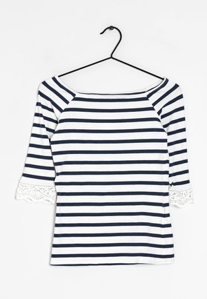 Laivastonsininen ja valkoinen vaakaraitainen kolmen neljänneksen hihan paita, jossa on pitsireunus hihoissa, mustalla vaateripustimella roikkuen valkoista taustaa vasten.