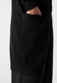 Pull noir en tricot avec des poignets côtelés et un détail de poche, présentant une texture douce et une coupe décontractée atteignant les hanches.