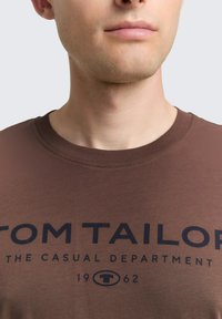Bruine katoenen t-shirt met ronde hals, voorzien van een zwart gedrukt logo "TOM TAILOR" en tekst eronder, zachte textuur en korte mouwen.