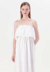 Hvit ermeles kjole med samlet liv og broderte blomsterdetaljer på det øverste laget, flytende skjørt og minimalistisk design.