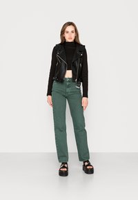 Colete de couro preto, estilo biker, por cima de um suéter preto de gola alta, combinado com jeans verdes de cintura alta e sandálias pretas de plataforma.