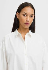 Camicia bianca con colletto classico, caratterizzata da una vestibilità comoda e una texture morbida. Orecchini a cerchio dorati aggiungono un accento sottile.