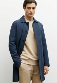 Granatowy płaszcz trench z kołnierzykiem, z przodu zapinany na guziki i z bocznymi kieszeniami, nałożony na beżowy sweter i dopasowane spodnie.