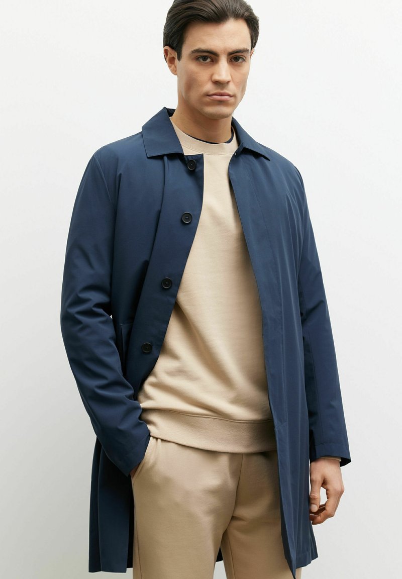 Granatowy płaszcz trench z kołnierzykiem, z przodu zapinany na guziki i z bocznymi kieszeniami, nałożony na beżowy sweter i dopasowane spodnie.