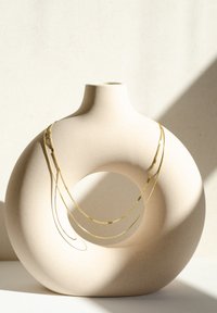 Goldene Ketten liegen über einem rundlichen, beigen Keramikaussteller mit einem zentralen Loch, der eine glatte, matte Oberfläche und subtile Schatten aufweist.