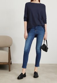 Pull en maille marine, manches trois-quarts ; jeans skinny bleus ; mocassins noirs ; petit sac à main noir avec poignée supérieure. Design minimaliste.