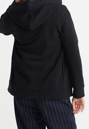 Person wearing a black hooded jacket and dark navy pants with white vertical pinstripes, shown from the back against a white background.