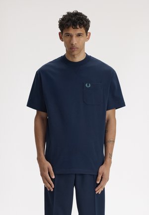 Mann trägt ein marineblaues Kurzarmhemd mit Brusttasche und kleinem Logo, steht vor einem einfarbigen hellen Hintergrund.