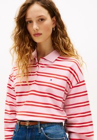 Ροζ μπλούζα πόλο με μακρύ μανίκι και κόκκινες οριζόντιες ρίγες, με γιακά και μικρό λογότυπο. Φορεμένη με ψηλόμεσο μπλε τζιν παντελόνι.