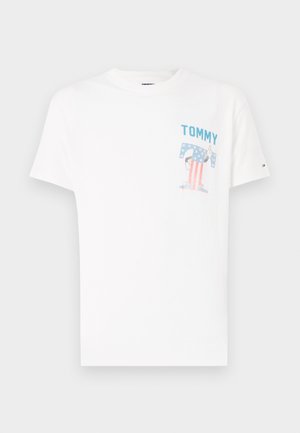 T-shirt bianca in cotone con la scritta blu "TOMMY" e un'immagine di un personaggio con un motivo a bandiera americana nella zona della tasca sul petto.