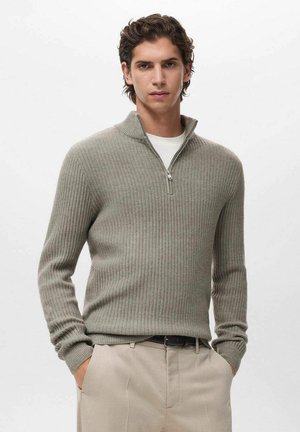 Suéter de rayas en gris claro con cuello con cremallera, hecho de un material suave. Lleva puesto sobre una camisa blanca, acompañado de unos pantalones beige claros.