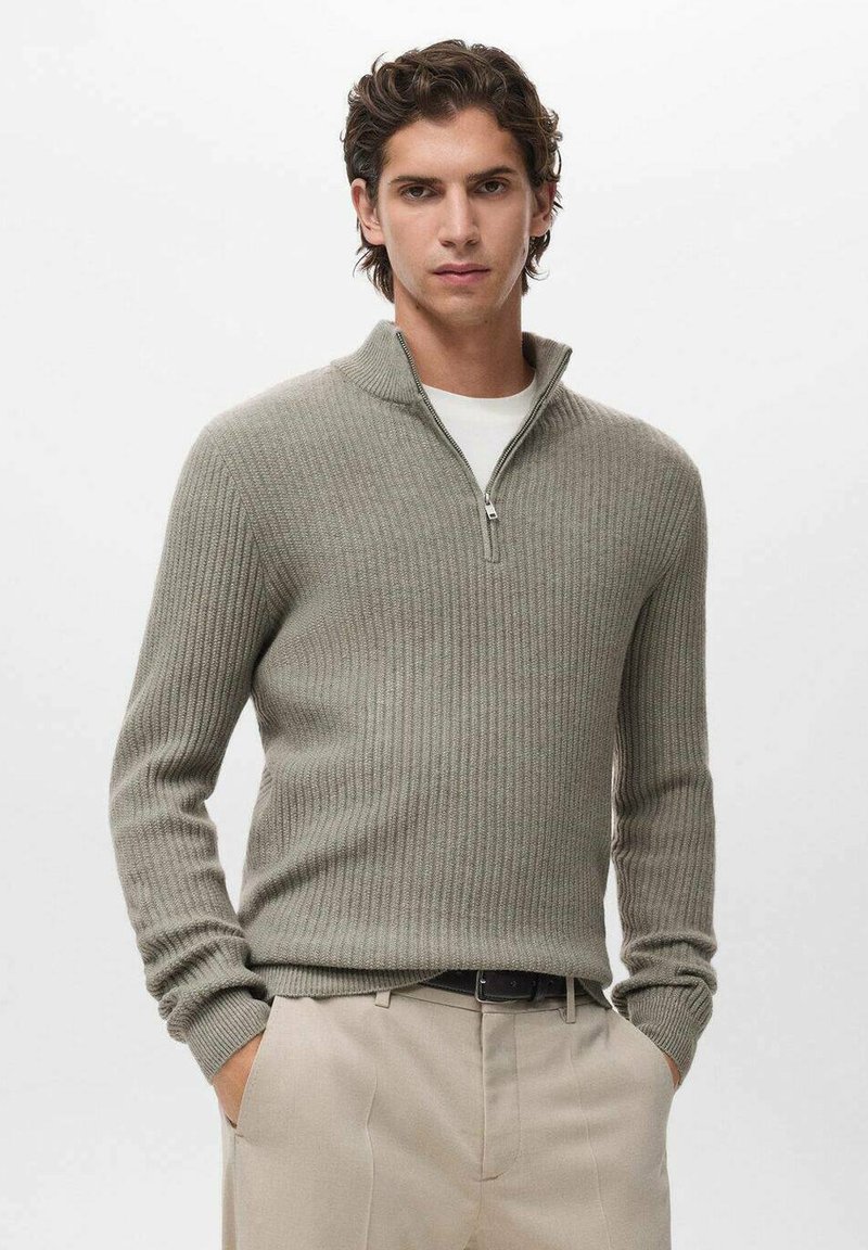 Pullover grigio chiaro a coste con colletto a zip, realizzato in un materiale morbido. Indossato sopra una maglietta bianca, abbinato a pantaloni beige chiaro.