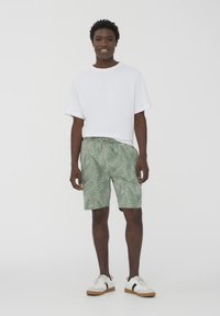 T-shirt bianca, shorts verdi con motivo fogliare, vita con coulisse, modello in piedi con sneakers bianche su uno sfondo neutro.