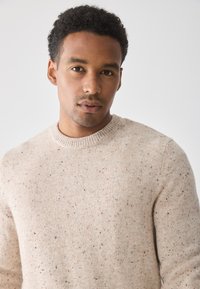 Beiger gestrickter Pullover mit Rundhalsausschnitt, der orangefarbene und schwarze Sprenkel aufweist. Die Stoffstruktur ist glatt, dicht und mit einem subtilen Muster versehen.