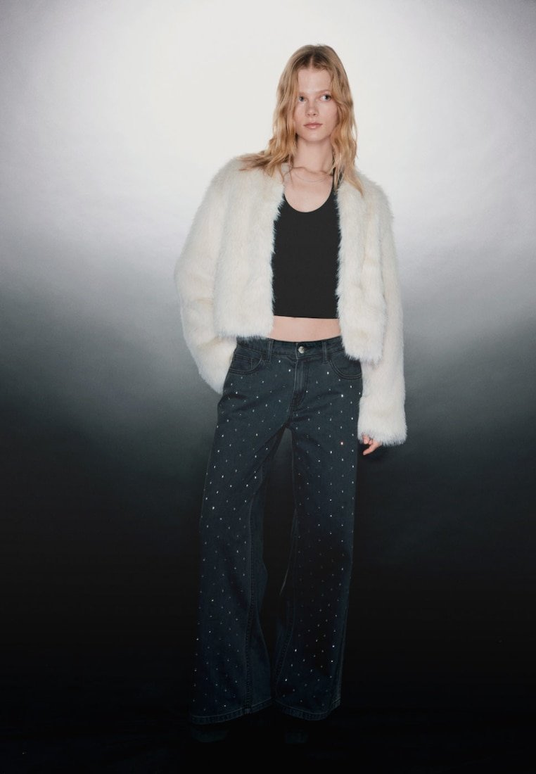 Chaqueta corta blanca y esponjosa, camiseta negra sin mangas, vaqueros de pierna ancha en denim oscuro con tachuelas plateadas, modelo posando contra un fondo degradado gris.