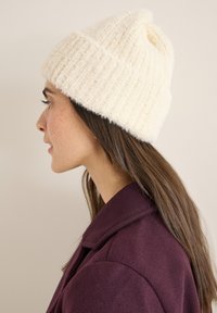 Beanie lavorato a maglia color crema con texture a coste e orlo ripiegato, indossato da una persona con capelli lunghi, abbinato a un cappotto di un viola scuro.