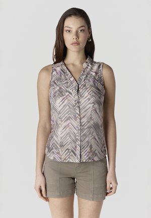 Giovane donna che indossa una camicetta smanicata con bottoni e motivo a fantasia, e pantaloncini color kaki, in piedi davanti a uno sfondo grigio chiaro uniforme.