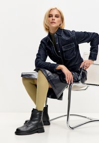 Stivali neri in pelle alla caviglia con cerniera, abbinati a collant verdi oliva con texture e a un vestito camicia di jeans con gonna nera lucida.