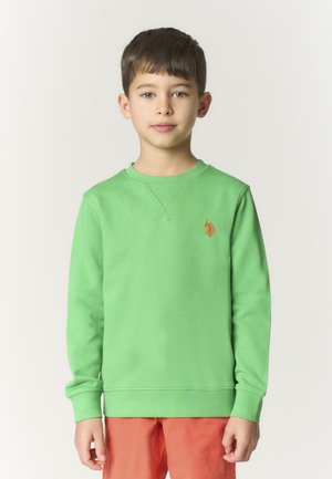 Ragazzo in piedi, rivolto in avanti, che indossa una felpa verde brillante con logo arancione e pantaloni color corallo su uno sfondo chiaro.