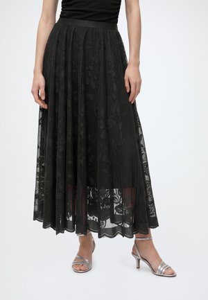 Mulher a usar uma saia midi preta de renda floral, um top preto simples e sandálias prateadas de salto alto com tiras, em fundo claro.