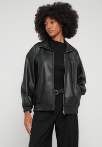 Jachetă neagră din piele ecologică, cu închidere cu fermoar, guler drept și manșete adunate. Are croială lejeră și textură fină.