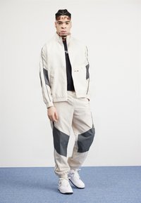 Light beige athletic tracksuit with gray accents, featuring a zip-up jacket and tapered pants, paired with white sneakers on a blue background.