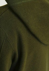 Olijfgroene hoodie gemaakt van zacht, textuurachtig breimateriaal. Bevat een kangaroezak en een aangepaste capuchon. Geen zichtbare hardware.