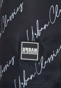 Fekete szövet, amelyen fehér, dőlt betűkkel a "Urban Classics" felirat található egy négyzet alakú címkén. Sima textúra és egyszerű dizájn.