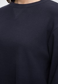 Tmavomodrý pulóver s dlhým rukávom a rebrovanou štruktúrou, s okrúhlym výstrihom a jemným trojuholníkovým detailom pri golieri.