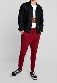 Svart denimjacka över en vit grafisk T-shirt, ihop med röda joggers med svarta sidolinjer och svarta sneakers med vita detaljer.