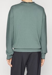 Personne portant un pull à manches longues turquoise avec le logo Hugo noir sur le col et un pantalon noir ample, vue de dos.