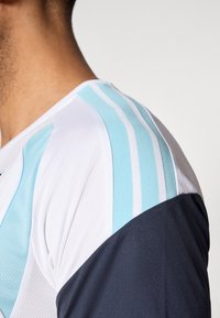 Αθλητική φανέλα με λευκό ύφασμα και ανοιχτόμπλε και σκούρο μπλε λεπτομέρειες, που ενσωματώνονται υφή διχτυού και ρίγες στους ώμους.