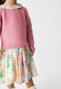 Vaaleanpunainen neulepaita, jossa on röyhelömäinen kukallinen kaulus, yhdistettynä polvipituiseen hameeseen, jossa on värikkäitä kukkakuoseja pehmeällä taustalla.